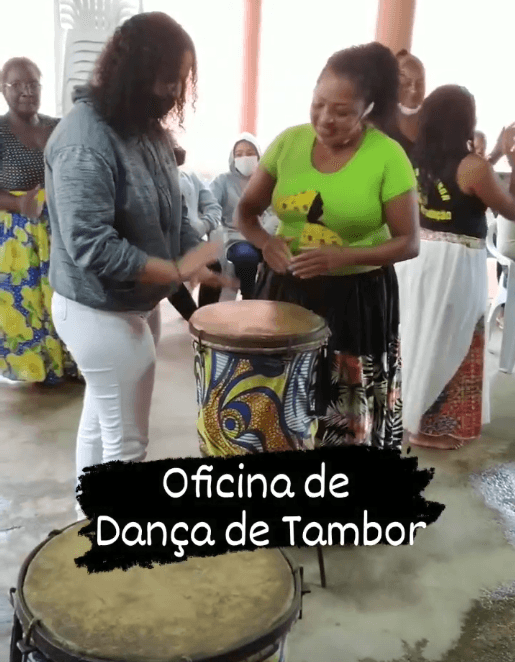Oficina de dança do tambor. A coordenadora de saberes Domingas Quilombola com outra mulher tocando um tambor em um espaço cultural.