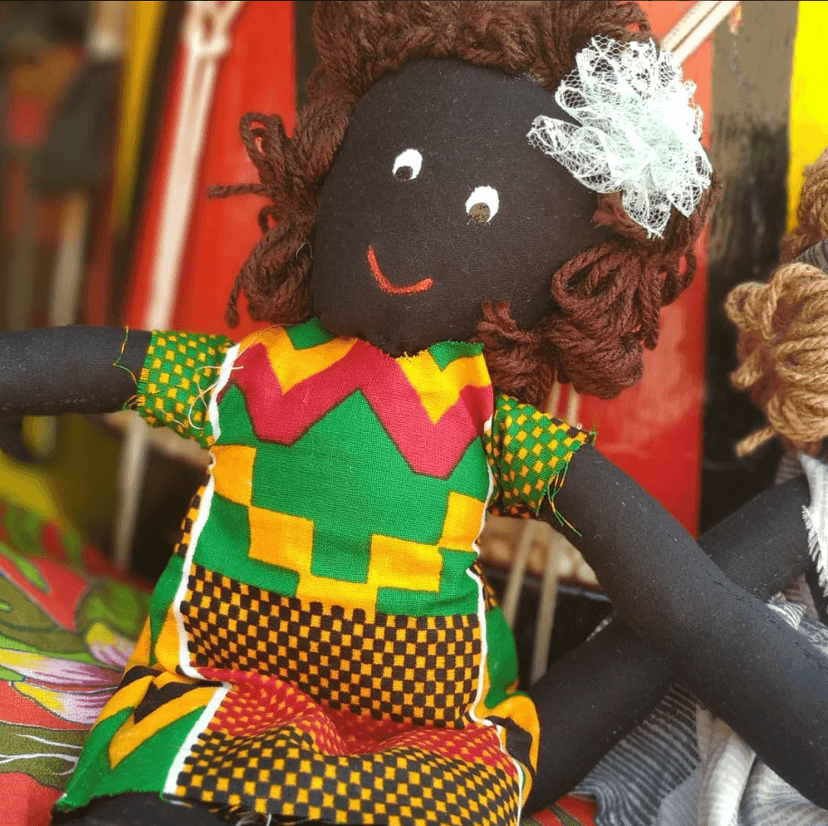 Boneca negra artesanal usando um enfeite branco no cabelo e vestida com um vestido colorido nas cores verde, vermelha, amarela, laranja e detalhes em preto.